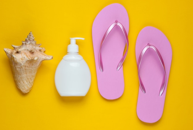 Beach accessories. Fashionable beach pink flip flops, sunblock bottle, seashell on yellow paper background.