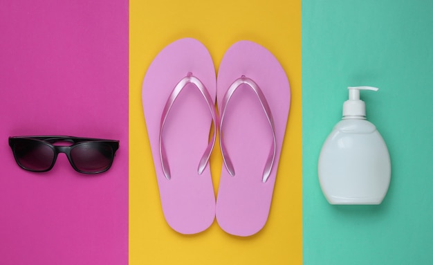 Пляжные аксессуары. Модные пляжные розовые шлепанцы, бутылка солнцезащитного крема, солнцезащитные очки на фоне цветной бумаги. Плоская планировка. Вид сверху