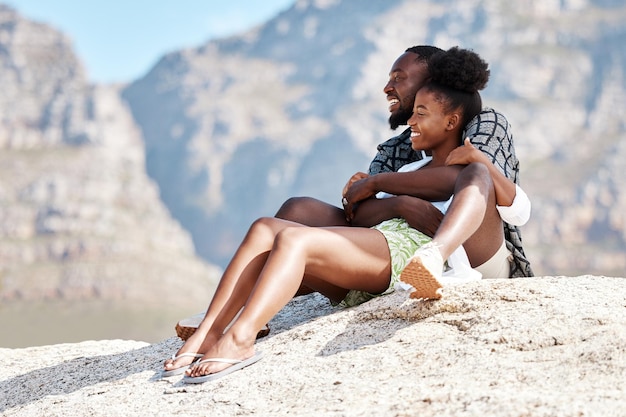 여름 휴가 여행에서 남아프리카 공화국 산을 배경으로 바위 위에 있는 해변 흑인 커플과 행복한 여성과 남성 미소는 사람들을 편안하게 하고 보안 신뢰 또는 안전 러브 본드에서 자연의 전망을 즐깁니다.