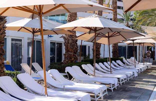 Beach chairs umbrellas around swimming pool with palm trees background Tourism summer concept Nice beach sun beds with a white towels Empty lounge chair in a row with umbrella under the sunlight