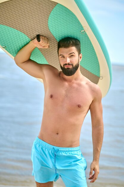 On a beach. A dark-haired bearded young man carrying kayak