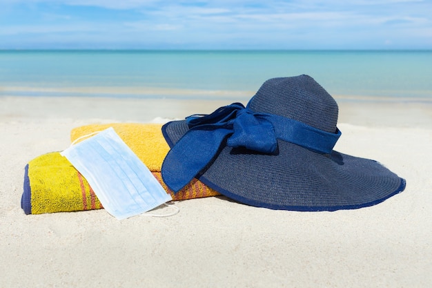 Cappello da spiaggia con asciugamani e mascherina medica sulla sabbia.