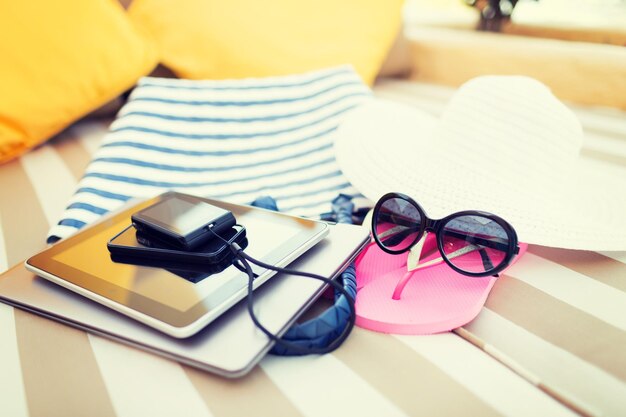 beach, summer vacation and technology concept - close up of tablet pc, notebook, smartphone and summer accessories on beach