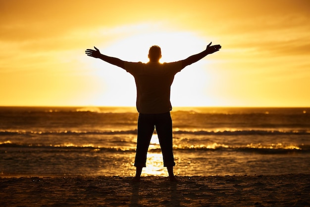 Beach sunset and silhouette of man in nature with arms to the sky carefree while on vacation Freedom shadow and guy standing outdoor by sea or ocean on holiday adventure or journey in Australia