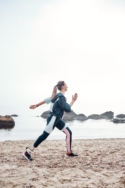 Beach workout vertical photo of strong sporty disabled woman with prosthetic leg is running on