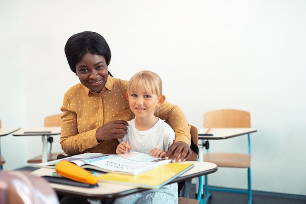 Beaming helpful teacher hugging cute blueeyed smart girl