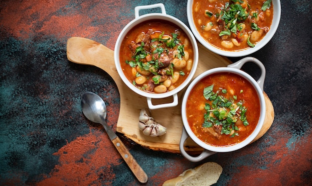 Фасолевый суп с мясом и овощами подается на деревенской доске с чесноком. Традиционный балканский суп или рагу Корбаст Пасулдж (Грах). Вид сверху, копировать пространство