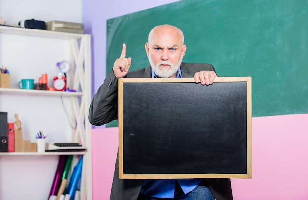 Bear in mind exam and graduation results information is here senior man teacher with empty blackboard place for copy space Mature teacher man hold chalk board back to school Education concept