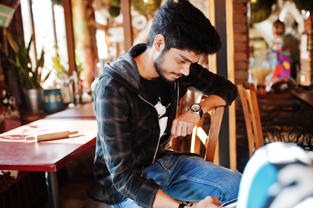 Бородатый азиат в повседневной одежде сидит в крытом кафе и смотрит на свой телефон