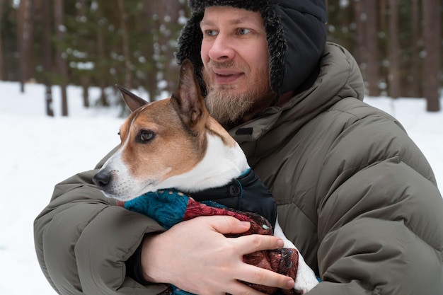 бородатый мужчина обнимает маленькую собачку, гуляющую в зимнем лесу