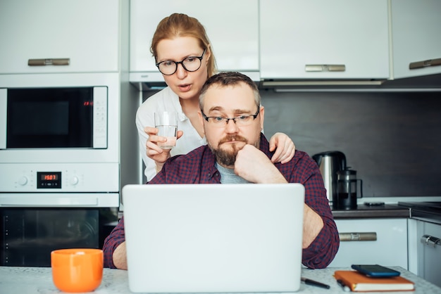 Beard man, and woman with glasses in kitchen looking at laptop, discussing the morning news. Adult couple spending time together at home.