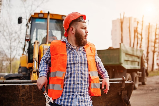 Beard worker man suit construction worker in safety orange helmet against tractor with adjustable wrench at hand