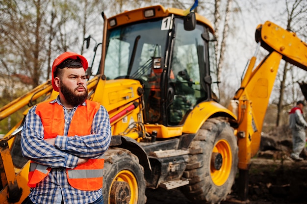 Beard worker man suit construction worker in safety orange helmet against tractor.