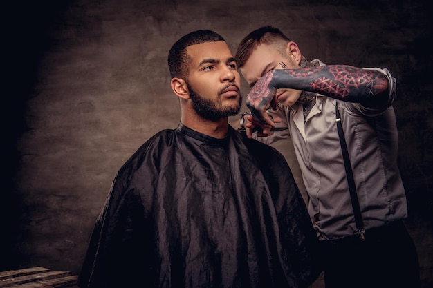 Bearded African American hipster guy getting a haircut by an old-fashioned tattoed professional hairdresser does the haircut.