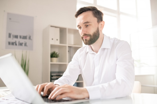 Foto analista o broker barbuto in camicia bianca che guarda il display del laptop durante la navigazione in rete e la ricerca di dati online