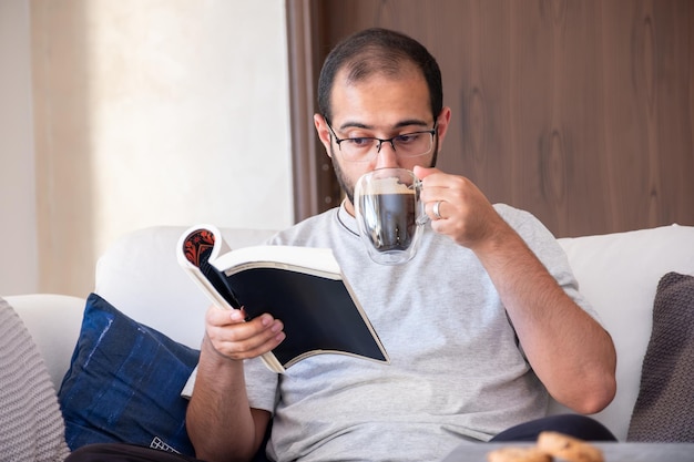 Bearded arab man drinking coffee while reading book and sitting on living room couch