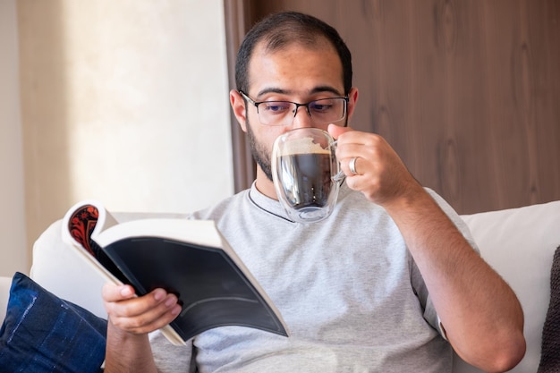 Foto uomo arabo barbuto che beve caffè mentre legge un libro e si siede sul divano del soggiorno