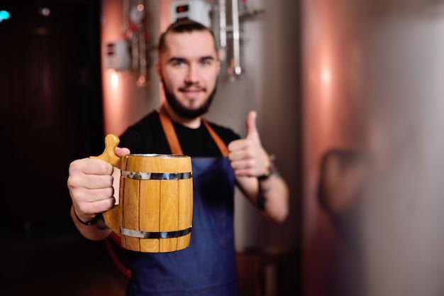 Bearded attractive male brewer with wooden beer mug in hand on background of brewery