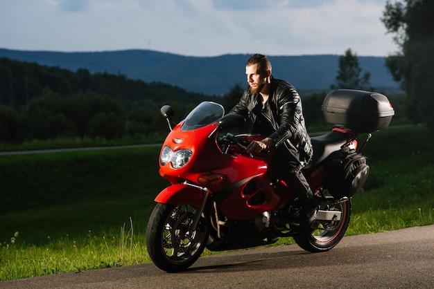 革のジャケットを着たひげを生やしたバイカーは、夕方に赤いスポーツバイクに乗る