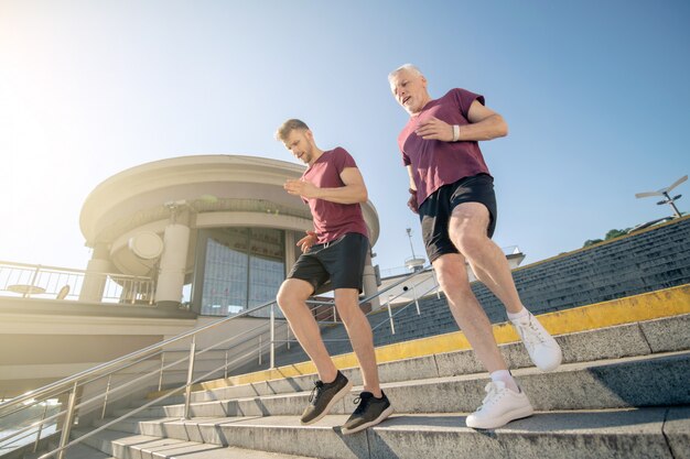 Photo bearded brown-haired male and mature grey-haired male running down stairs