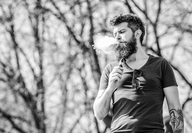 Бородатый брутальный мужчина, курящий электронную сигарету, хипстер, мужчина держит устройство для вейпинга, безопасность для здоровья и наркомания, вдыхание пара, мужчина, курящий электронную сигарету, зрелый хипстер с бородой, такой вкусный