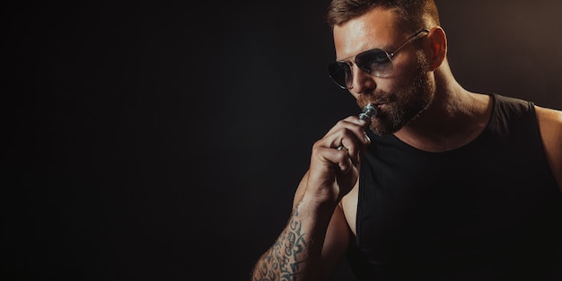 Bearded brutal male in sunglasses smoking a vapor cigarette as an alternative to tobacco on black