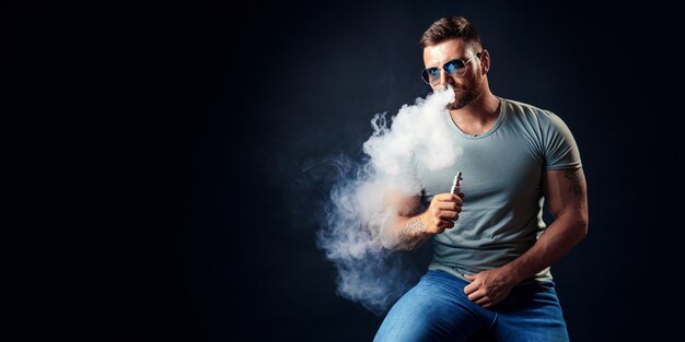 Bearded brutal male in sunglasses smoking a vapor cigarette as an alternative to tobacco studio shot on black 