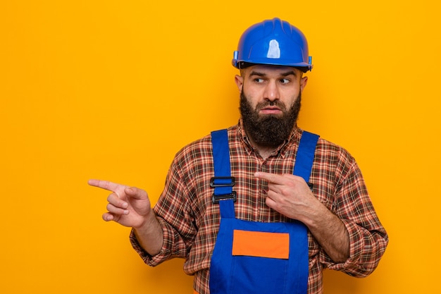 Bearded builder man in construction uniform and safety helmet looking aside with serious face pointing with index fingers to the side standing over orange background