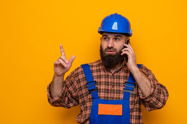 Photo bearded builder man in construction uniform and safety helmet looking aside with serious face showing index finger while talking on mobile phone