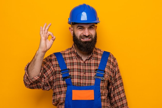 Bearded builder man in construction uniform and safety helmet looking at camera smiling cheerfully doing ok sign standing over orange background