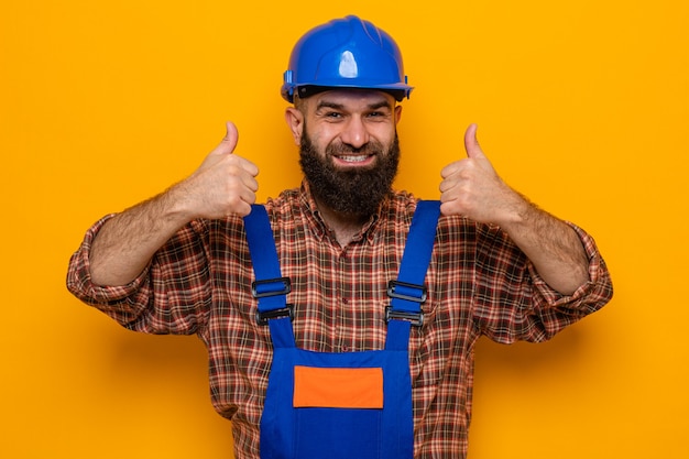 Uomo barbuto costruttore in uniforme da costruzione e casco di sicurezza che sembra felice e allegro sorridente che mostra i pollici in su