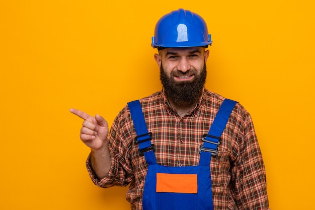 Bearded builder man in construction uniform and safety helmet looking happy and positive pointing with index finger to the side smiling