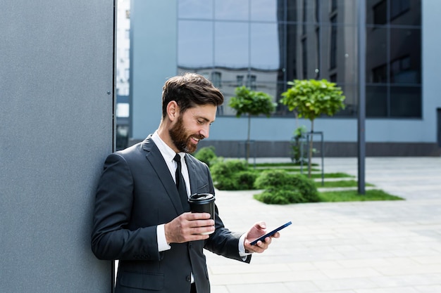 Bearded businessman in formal suit on break using mobile phone use smartphone. business man standing outside on modern urban city street background with coffee cup in downtown outdoors. copy space