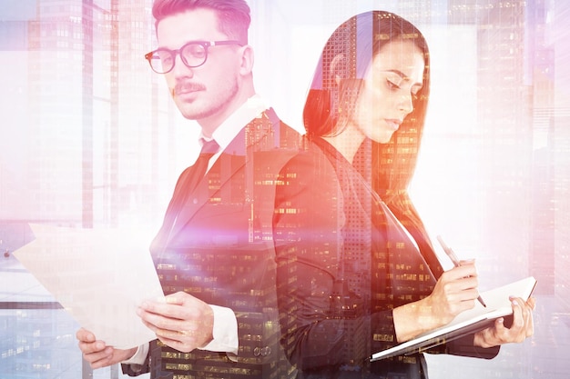 Bearded businessman in glasses reading document standing near serious businesswoman taking notes with double exposure of night city. Toned image