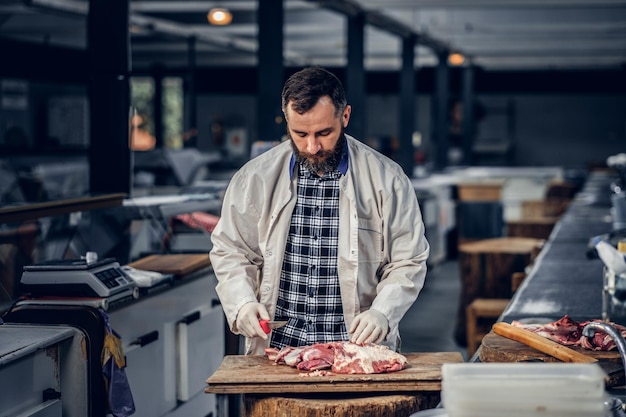 Bearded butcher dressed in a fleece shirt cut fresh bacon meat.