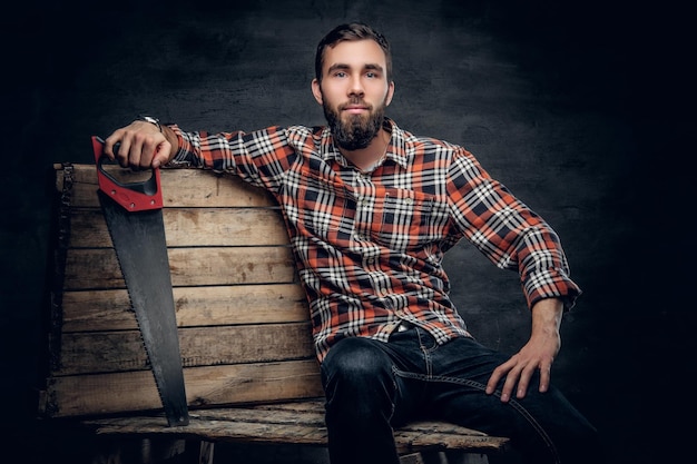 Photo a bearded carpenter male dressed in a jeans and a plaid shirt gits on a wooden palette and holds handsaw.