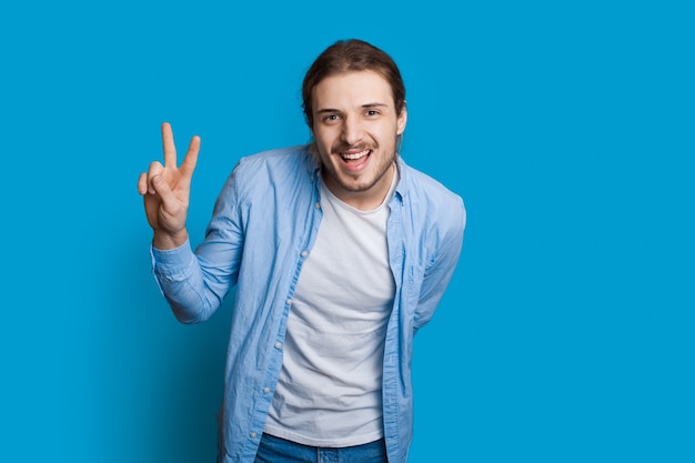 Bearded caucasian man is gesturing the hello and peace sign smiling at camera on a blue wall in casual clothes