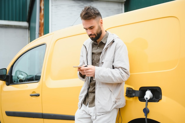 Bearded caucasian man standing near an electric car that is charging and making time adjustments on a smartphone