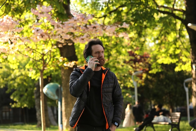 A bearded Caucasian man talking via smartphone in front of a blurry background with soft natural lig