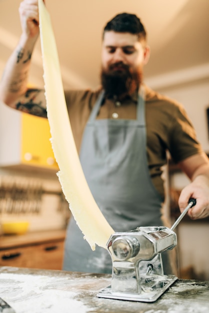 Photo bearded chef in apron works with dough in pasta machine on wooden kitchen table. homemade spaghetti cooking process