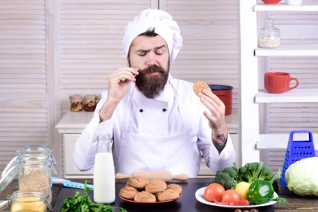 Bearded chef hold oatmeal cookies. Handsome chef, cook or baker tastes oatmeal cookies. Professional cook in white suit in kitchen. Green grocery concept. Organic food. Dairy products, cooking concept