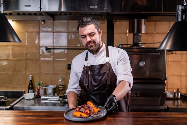 Bearded chef in leather apron putting prepared dish on counter in restaurant