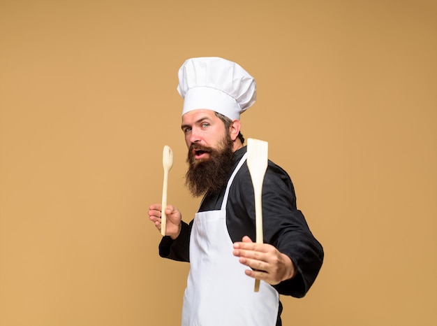 Cuoco barbuto con cucchiaio di legno e spatola in mano capo uomo in uniforme da cuoco tiene cucina in legno