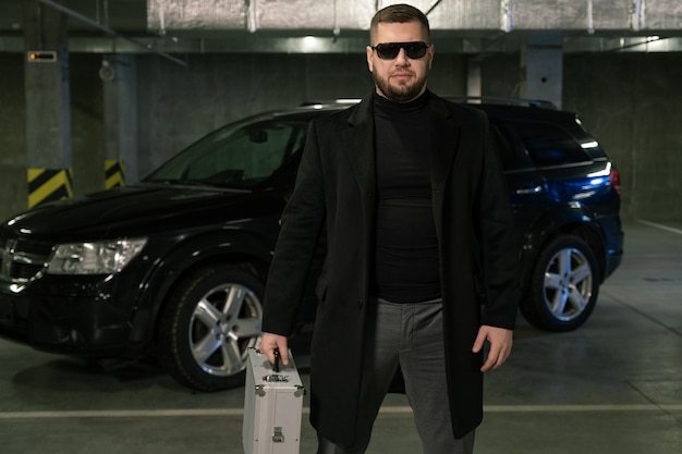 Bearded criminal or agent in black coat and sunglasses moving down parking area while carrying suitcase with cash or drugs