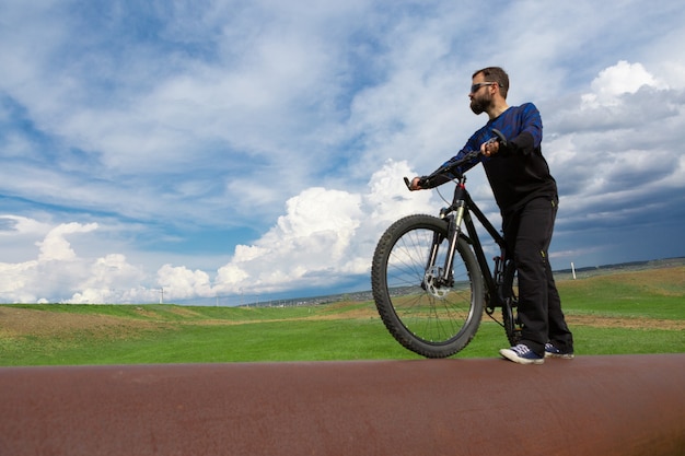 Бородатый велосипедист на горном велосипеде на ржавой трубе зеленая трава голубое небо