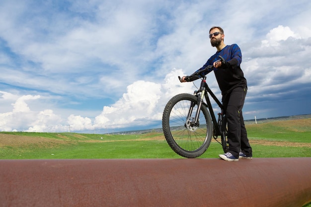 Bearded cyclist on a mountain bike on a rusty pipe green grass blue sky