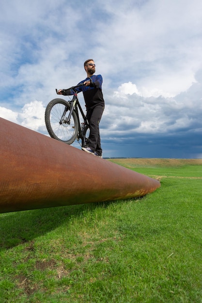 Ciclista barbuto su una mountain bike su un tubo arrugginito erba verde cielo blu