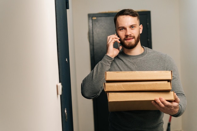 Bearded delivery man standing by door of customer apartment with boxes of hot pizza and calling customer on phone looking at camera