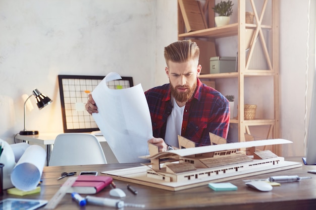 Bearded designer sitting at the table and holding drawing of wooden model of building. Engineering and architectural concept