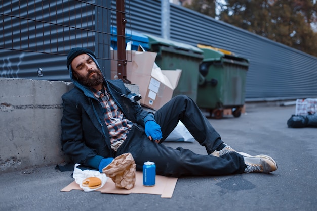 Bearded dirty beggar with food sitting at the trashcan on city street.